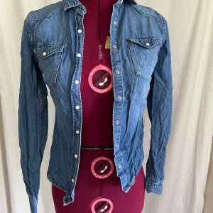 Jeansskjorta från Crocker XXS - Snygg jeansskjorta från Crocker i klassisk blå denim. Skjortan har långa ärmar, tryckknappar framtill och två bröstfickor med lock. Perfekt för dig som gillar en tidlös och avslappnad stil.