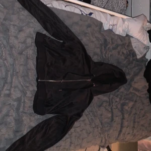 Svart hoodiejacka med dragkedja - Svart hoodiejacka i mjukt material med hel dragkedja framtill och justerbar huva. Jackan har långa ärmar och en avslappnad passform, perfekt för chill dagar eller när du vill ha en enkel streetstyle-look.