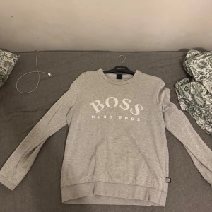 Grå sweatshirt från Hugo Boss L - Snygg ljusgrå sweatshirt från Hugo Boss i storlek L. Tröjan har klassisk rund hals, långa ärmar och stor BOSS-logga framtill. Tillverkad i mjuk bomullsmix som är skön att ha på sig. Perfekt för en chill och stilren look.