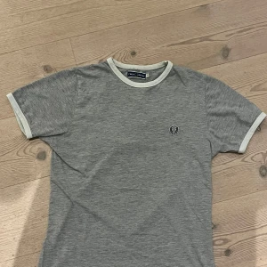 Grå t-shirt från Fred Perry - Ruggigt snygg grå t-shirt från Fred Perry med vit ribbad kant vid hals och ärmslut. Klassisk logga broderad på bröstet. Skön passform och stilren design, perfekt till jeans eller shorts. Tröjan är i skick! Bara att höra av sig vid eventuella frågor!😉