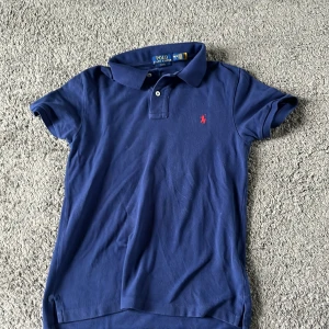 Marinblå pikétröja Polo Ralph Lauren - Snygg marinblå pikétröja från Polo Ralph Lauren i slim fit-modell. Tröjan har klassisk krage, korta ärmar och två knappar framtill. Ikoniska röda broderade loggan på bröstet. Tillverkad i mjuk bomull som är skön mot huden.
