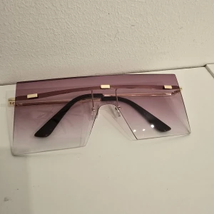 Oversized lila solglasögon med guld - Stora, futuristiska solglasögon med lila tonade glas och gulddetaljer upptill. Raka linjer och shield-form ger en cool vibe. Svarta skalmar och transparent nederdel på glasen. Perfekta för dig som vill sticka ut.