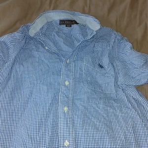 Blårutig skjorta från U.S. Polo Assn. - Snygg blårutig skjorta från U.S. Polo Assn. i 100% bomull. Klassisk krage, kortärmad och med knappar framtill. Perfekt för dig som gillar en stilren och fräsch look med en sportig touch.