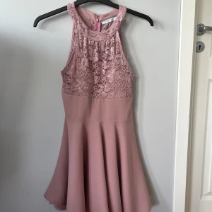 Rosa spetsklänning med utställd kjol - Supersöt rosa kortklänning med spets upptill och slät, utställd kjol. Hög halsringning och ärmlös design. Dragkedja bak för enkel påklädning. Det står tyvärr ingen storlek i men skulle gissa på ca 36.