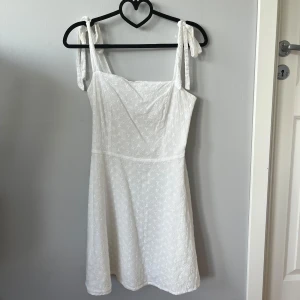 Vit kort klänning - Superfin vit kort klänning med knytbara axelband och broderade små blommor. Klänningen har en lätt utsvängd kjol. En väldigt fin och söt klänning!