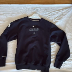 Svart Lakers Nike Dri-Fit tröja - Svart crewneck tröja från Nike med Lakers-tryck på bröstet och NBA-logga på ärmen. Tillverkad i Dri-Fit material som andas, perfekt för basket eller chill. Klassisk passform med långa ärmar och ribbade muddar. Snygg och sportig vibe.