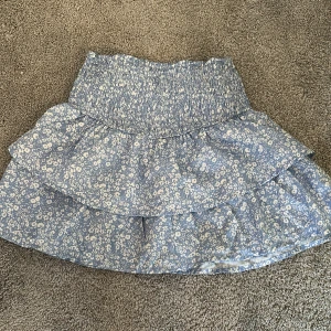 Blå blommig volangkjol - Supersöt blå kortkjol med vita små blommor och bred smock i midjan. Kjolen har dubbla volanger som ger en flowy och lekfull känsla. Perfekt för varma dagar och lätt att matcha med en enkel topp.