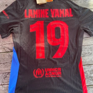 FC Barcelona Lamine Yamal Nike tröja - Snygg FC Barcelona fotbollströja med Lamine Yamal och nummer 19 på ryggen. Tröjan är svart med röda och blå detaljer, officiella klubbmärket och Nike-logga framtill. Kortärmad modell i lätt Dri-FIT material, Champions League-märke på ärmen och Spotify-logga på bröstet.