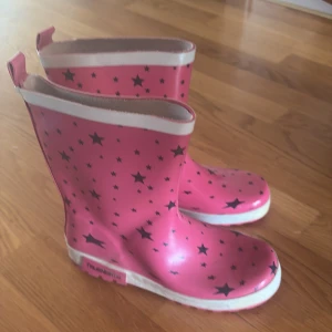Rosa gummistövlar med stjärnor - Coola rosa gummistövlar med svarta stjärnor och vit kant upptill. Perfekta för regniga dagar och festivaler. Tillverkade i syntetmaterial som håller fötterna torra. Snygg detalj med dragflik bak för enkel påtagning.