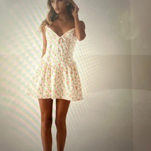 Peppermayo  - Superfin kortklänning i ljusbeige med små rosa blommor, v-ringning och smickrande snörning framtill. Klänningen har breda axelband och en lätt utställd kjol som ger en romantisk vibe. Perfekt för varma dagar och somriga tillfällen!