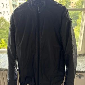 Svart dunjacka från Houdini, herr L - Svart dunjacka från Houdini i herrmodell, storlek L. Jackan är lätt och smidig med dragkedja framtill och huva. Tillverkad i syntetmaterial med Primaloft-foder som håller dig varm utan att bli klumpig. Perfekt för kyliga dagar och outdoor-häng.