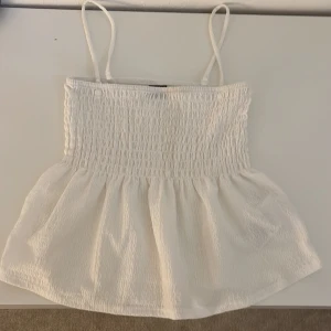 Vit smockad linnetopp Gina Tricot - Supersöt vit linnetopp från Gina Tricot i storlek XS. Toppen har smala axelband, smockad överdel och volangig nederdel. Perfekt för varma dagar och ger en fräsch och luftig känsla. Materialet är lätt och har en diskret struktur.