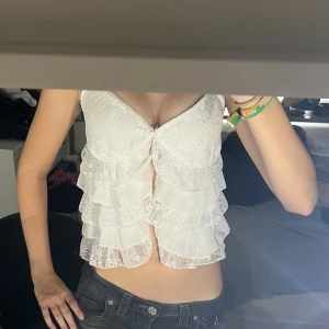 Vit spetsig volangblus med knappar - Supersöt vit blus i spets med flera lager volanger och v-ringning. Toppen har smala axelband och knäppning framtill med små knappar. Spetsen ger en romantisk vibe och volangerna gör den extra unik och lekfull. Man måste knyta snöret runt knappen för snöret har gått upp! Annars toppen skick 