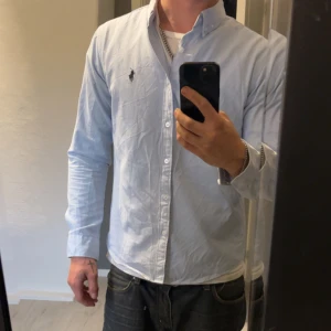 Ljusblå Polo skjorta  - Klassisk ljusblå snygg Polo skjorta med button down-krage och broderad logga på bröstet. Skjortan är tillverkad i mjuk bomull, använd 3 gånger och är perfekt för dig som gillar stilrena och tidlösa plagg.