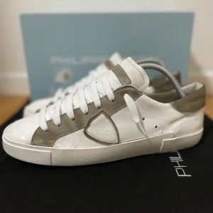 Philippe Model vita sneakers - Snygga vita sneakers från Philippe Model med grågröna mockadetaljer och klassisk logga på sidan. Skorna har rund tå, platt sula och vita snören. Tillverkade i skinn och mocka för en lyxig känsla. Perfekta för dig som gillar stilrena och trendiga sneakers. Storlek 42, Bra skick! Orginalbox medföljer! Nypris runt 3500 kr!!! Sulan är proffesionellt reparerad av skomakare. Hör av er ifall ni har några frågor! 🙌