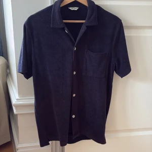 Mörkblå frottéskjorta  - Säljer en mörkblå skjorta i mjuk frotté. Skjortan har korta ärmar, klassisk krage, bröstficka och knäppning framtill med vita knappar. Perfekt för en avslappnad och stilren look.