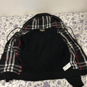 Svart hoodie med dragkedja  Burberry - Svart hoodie med dragkedja och klassiskt Burberry-rutigt foder i huvan. Jackan har fickor framtill och justerbar huva med snörning. Perfekt för en stilren och avslappnad look.
