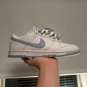 Nike Dunk Low i vitt och ljusblått - Nike Dunk Low sneakers i vitt skinn med ljusblå detaljer och sula. Skon har klassisk snörning och perforerad tå för extra ventilation. Perfekt för dig som gillar stilrena och sportiga sneakers. Andvända fåtal gånger 