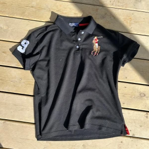 Svart pikétröja från Polo Ralph Lauren - Säljer en svart pikétröja från Polo Ralph Lauren med klassisk polospelare broderad på bröstet och en stor vit siffra 9 på ärmen. Tröjan har krage, knappar och röda detaljer på insidan av kragen. Upplevs som xs-s storleken, åris går diskuteras. 