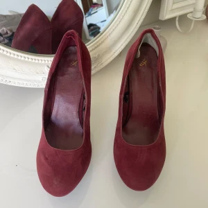 Vinröda pumps med klack - Säljer ett par vinröda pumps i mocka med hög klack . Skorna har en elegant och klassisk design som passar perfekt till dig som vill sticka ut med färg. Snygga att matcha med både jeans och klänning.