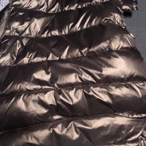 Svart dunväst från Moncler - Snygg svart dunväst från Moncler med glansig finish och två dragkedjefickor framtill. Västen har hög krage och Moncler-logga på bröstet. Perfekt för lager-på-lager under kyliga dagar. Lite sönder nere vid västen vilket gör att jag gått ner i pris köpt som present men är en äkta vara som man kan scanna och dubbelkolla själv i t.ex NK eller en Moncler affär 