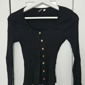 Svart ribbad kofta med träknappar - Svart ribbad kofta från H&M med långa ärmar och träfärgade knappar framtill. Tröjan har en tight passform och djup rundringning. Perfekt att styla till olika outfits.