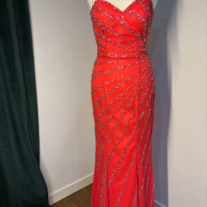 Röd glittrig långklänning med paljetter - Säljer en röd långklänning med glittriga paljettdetaljer i diagonala mönster. Klänningen är axelbandslös med en hjärtformad urringning och figurnära passform. Perfekt för bal eller festliga tillfällen.