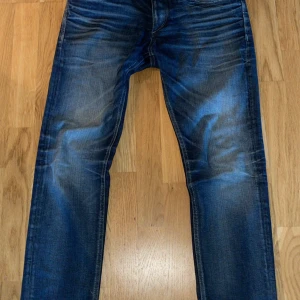 Blå jeans från Jack & Jones - Säljer ett par klassiska blå jeans från Jack & Jones med snygga slitningar och raka ben. Jeansen har fem fickor, normal passform och är tillverkade i denim. Perfekta för dig som gillar en tidlös stil.