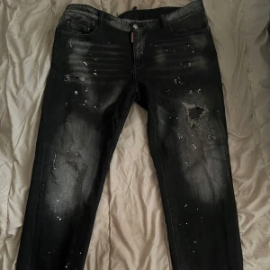 Svarta slim jeans från Dsquared2 med slitningar - Svarta jeans från Dsquared2 i slim fit-modell med coola slitningar och vita färgstänk. Jeansen har klassisk femficksdesign, knappgylf och unika detaljer som ger en trendig look.