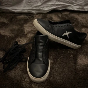 Svarta sneakers med vit fågelbrodyr - Säljer ett par svarta sneakers med vit broderad fågel på sidan. Skorna har vit platt sula och rund tå. Klassisk design utan synliga märkesloggor, passar till många stilar. Skorna är i syntetmaterial och har snörning,snörena snöras på vid köp! Axel arigato 