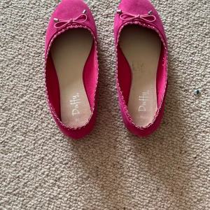 Rosa ballerinaskor med rosett från Duffy - Säljer ett par söta rosa ballerinaskor från Duffy med dekorativ rosett framtill och små silvriga detaljer. Skorna har rund tå och en tunn platt sula. Perfekta för dig som vill ha en färgklick till outfiten!