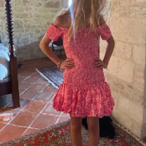 Rosa offshoulder kortklänning med volang - Wooooow säljer denna så populära rosa kortklänning med vitt mönster och offshoulder-modell. Klänningen har volang nertill och smockad överdel som ger en tight passform upptill. Perfekt för sommarens tillfällen! Endast använd 1 gång!