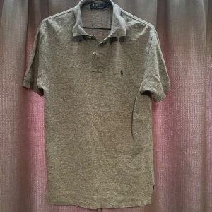 Pikétröja från Polo Ralph Laurent  - Klassisk grå pikétröja från Polo Ralph Lauren med korta ärmar och krage. Tröjan har två knappar vid halsen och den ikoniska broderade loggan på bröstet. Perfekt för en stilren och avslappnad look.