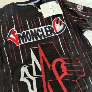 Svart t-shirt med Moncler-logga och ränder - Cool svart t-shirt från Moncler med stora broderade loggor och grafiska detaljer framtill. T-shirten har röda, vita och grå vertikala ränder samt rund halsringning. Märkespatch på ärmen och tryckt logga på ryggen.