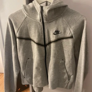 Grå zip-hoodie från Nike - Snygg grå hoodie från Nike med dragkedja och svart logga på bröstet. Jackan har huva, svarta detaljer och två fickor framtill. Perfekt för en sportig och avslappnad stil.