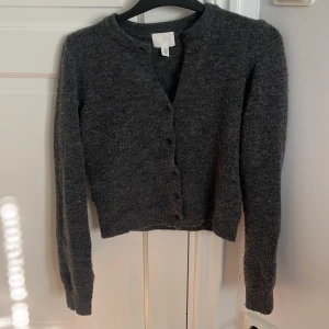 Mörkgrå stickad kofta från H&M - Mysig mörkgrå stickad kofta från H&M med rund halsringning och knappar framtill. Tröjan är croppad och har långa ärmar, perfekt för lager-på-lager under kyliga dagar.