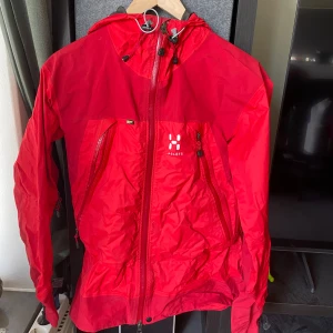 Röd skaljacka från Haglöfs - Röd skaljacka från Haglöfs med huva och flera dragkedjeförsedda fickor. Jackan har justerbar huva och är tillverkad i ett lätt syntetmaterial som passar perfekt för utomhusaktiviteter. Snygg och funktionell design med logga på bröstet.