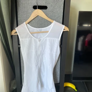 Vit ärmlös y2k topp - Säljer en vit ärmlös träningstopp i stretchigt material med dragkedja framtill. Toppen har rund halsringning och är tillverkad i en blandning av polyester och spandex för en bekväm passform. Perfekt för gymmet eller löpning.