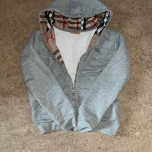 Grå hoodie med dragkedja från Burberry -  Säljer en helt ny Grå hoodie från Burberry med dragkedja och klassiskt rutigt foder i huvan. Mjukt material och ribbade muddar. Perfekt för en avslappnad och stilren look. De är bara och höra av er vid frågor. Pris kan sänkas lite vid snabb affär🤩