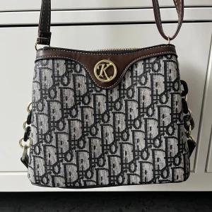 Brun och svart axelväska med monogram - Snygg axelväska med brunt och svart monogrammönster, guldfärgade detaljer och två dragkedjefack. Väskan har både axelrem och handtag, samt dekorativt K-emblem framtill. Vet tyvärr inte vad väskan är ifrån då jag köpte den privat. 