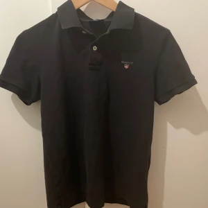 Svart pikétröja från Gant - Klassisk svart pikétröja från Gant i slim fit med korta ärmar och broderad logga på bröstet. Tröjan har krage och knappar framtill. Perfekt för en stilren och enkel look, den är skön och har ett skick på 9/10 med inga defekter. Slim fit