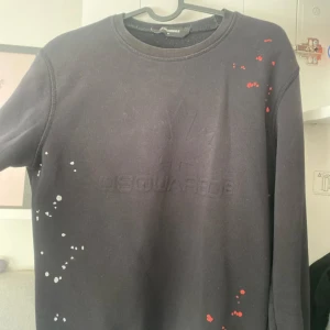 Svart sweatshirt från Dsquared2 - Svart sweatshirt från Dsquared2 med diskret broderad logga och lönnlöv på bröstet. Tröjan har rund halsringning och långa ärmar. Unika färgstänk i vitt och rött ger en cool touch till looken.