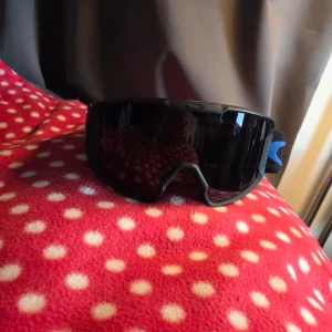 Oakley Prizm svarta sportglasögon - Säljer ett par svarta Oakley Prizm sportglasögon med bred, justerbar blå och svart rem. Glasögonen har en stor, rektangulär lins och är perfekta för utomhusaktiviteter som träning eller cykling.