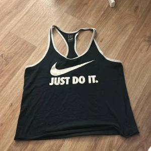 Svart linnetopp  - Säljer en svart linnetopp från nike med vitt tryck och texten 'Just Do It.' samt Nike-loggan på bröstet. Toppen har vita kantband och brottarrygg. Perfekt för träning eller sportiga outfits!