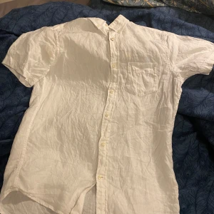 Vit linneskjorta från GANT - Säljer en klassisk vit kortärmad skjorta från GANT i linne. Skjortan har knappar framtill, bröstficka med broderad logga och en avslappnad passform. Perfekt för varma dagar och enkel att styla till olika outfits. Galet snygg till sommaren!