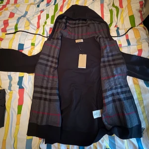 Svart hoodie från Burberry med rutig insida - Säljer en svart hoodiejacka från Burberry med klassiskt grått och rött rutigt foder på insidan. Jackan har dragkedja framtill och huva. T