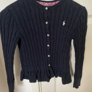 Polo Ralph Lauren stickad kofta - Mörkblå kabelstickad kofta från Polo Ralph Lauren med volangkant nedtill och vita knappar framtill. Klassisk rund halsringning och långärmad modell. Liten rosa logga broderad på bröstet. (Kids Size M!!! Men är som en normal Xs i vanlig storlek)