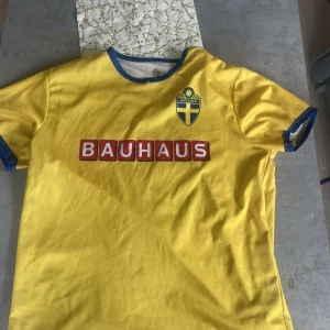 Svensk landslagströja med Bauhaus-tryck - Gul fotbollströja från svenska landslaget med blå detaljer vid krage och ärmslut. Bauhaus-logga på bröstet och ryggen samt nummer 22 i blått med vit kant. Broderad svensk landslagssköld på bröstet.
