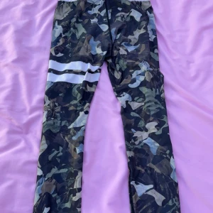 Camo leggings från Stronger - Säljer ett par camo-mönstrade leggings från Stronger med vita detaljer och bred midja. Perfekta för träning och har en snygg passform som sitter tight. Mjukt och stretchigt material för maximal komfort.