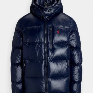 Mörkblå dunjacka från Polo Ralph Lauren - Säljer en mörkblå dunjacka från Polo Ralph Lauren med huva och dragkedja. Jackan har en klassisk pufferstil med broderad logga på bröstet och flera fickor med dragkedja. Perfekt för kyliga dagar.